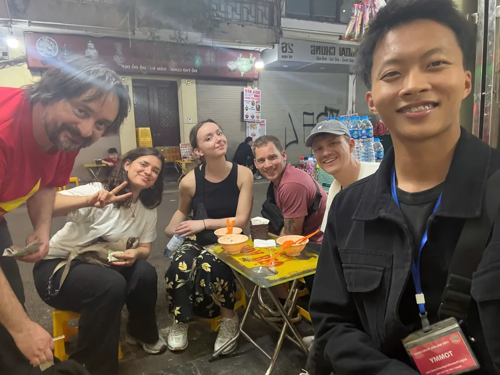 Small group seated at a low sidewalk table with bowls on an evening food walk.