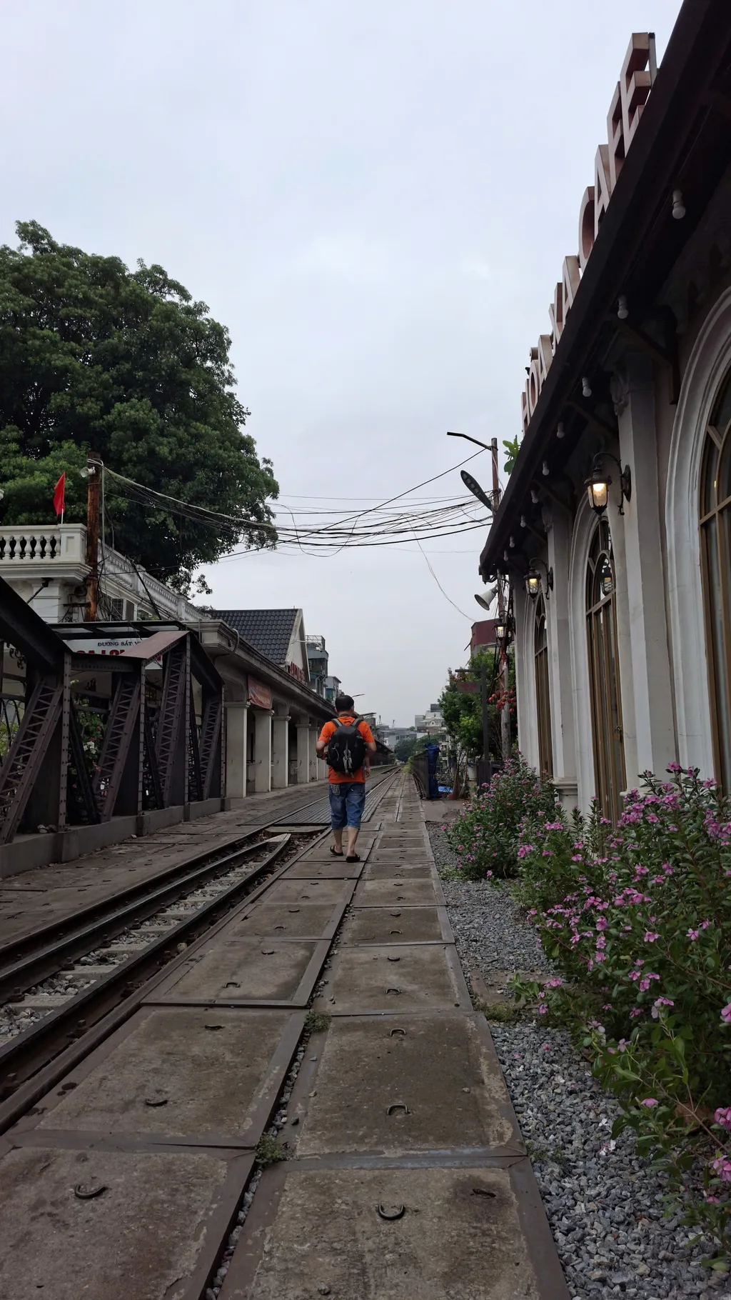 Active railway tracks beside a rusted metal truss and café frontage on a Hanoi rail corridor.